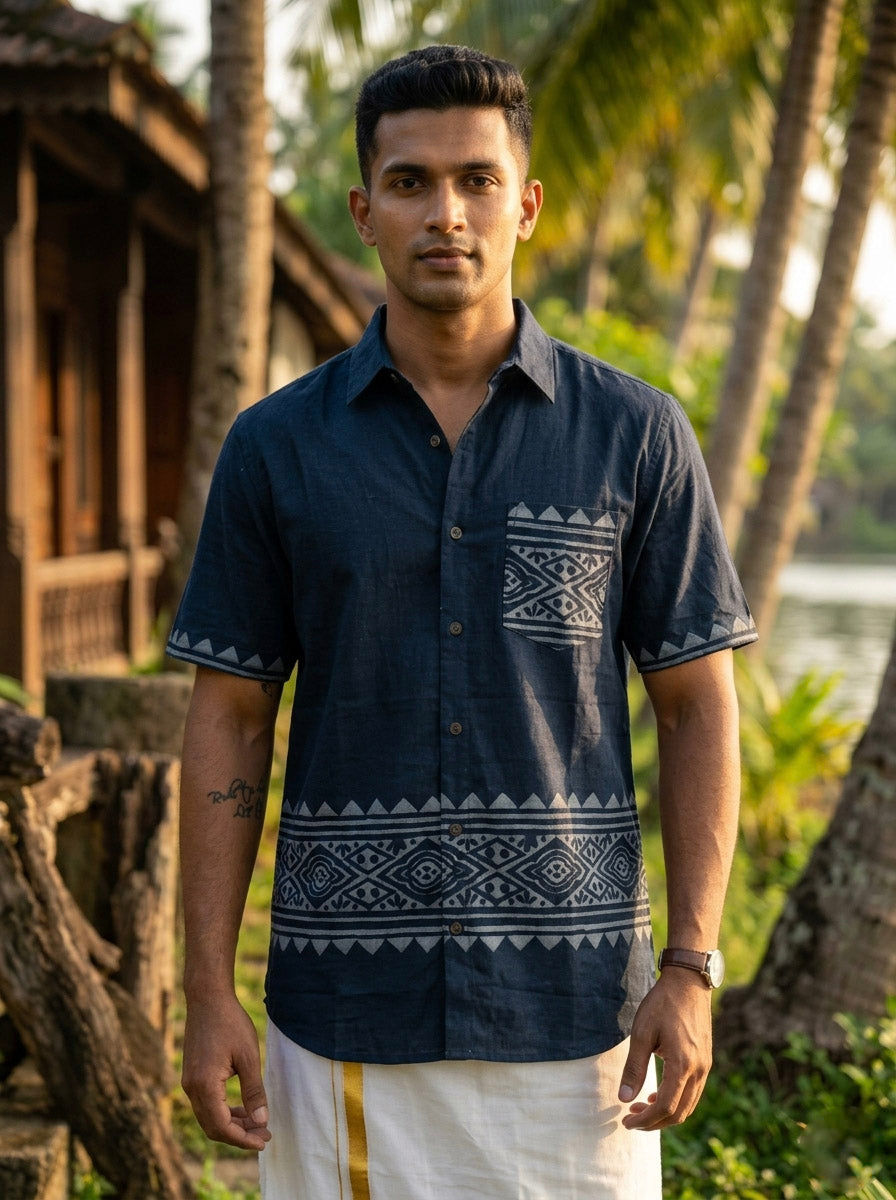 A man wearing a Bohemian Tribal Shirt in dark blue, featuring tribal block prints and crafted from breathable cotton fabric, pairs it with a white dhoti as he stands outdoors near palm trees and a wooden structure, looking at the camera.
