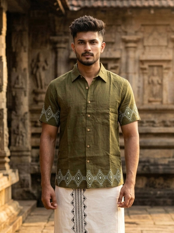Wearing the Vana Shirt in olive green, featuring white geometric patterns, paired with a classic white traditional garment, the man stands in front of an old carved stone building. The stylish bohemian Vana Shirt adds a contemporary touch to his timeless look.