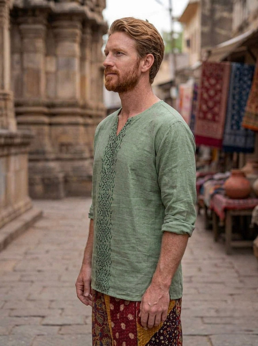 A man with red hair and a beard stands in a historic market street, wearing the light green Noma Shipibo Kurta paired with colorful patterned pants. Stalls with textiles and pottery are visible in the background.