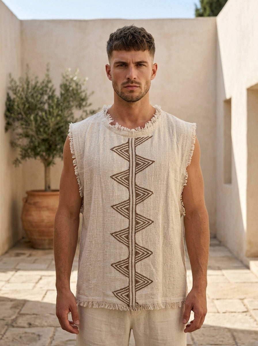 A man stands outside on a stone patio, wearing the Kosha Vest in Natural, a sleeveless garment featuring geometric patterns. Crafted from soft viscose knit, the vest exudes relaxed elegance. A large potted plant and cream-colored walls in the background enhance the scene’s laid-back atmosphere.