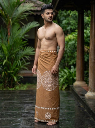 A barefoot man stands outdoors on wet stone, wearing the brown Kavu Dhoti with white mandala motifs. Surrounded by green plants and wooden pillars, he is shirtless and gazes to his left.