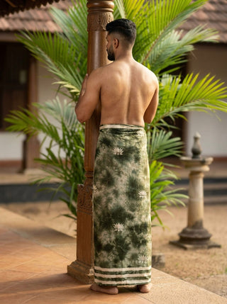 A man with short hair stands barefoot, back facing the camera, wearing the Lotus Shibori Dhoti next to a wooden pillar, with tropical plants and traditional architecture in the background.