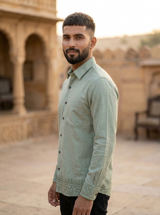 A man with short dark hair and a beard stands sideways outdoors, wearing the Madera shirt—a light green cotton design—paired with black pants. The background features stone architecture and soft, warm lighting.