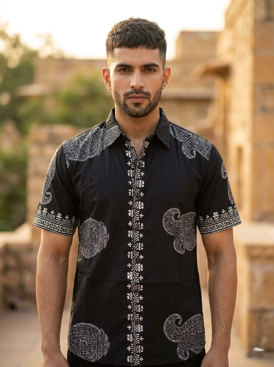 A man with short hair and a beard stands outdoors in front of rustic stone buildings and greenery, wearing the Obsid Shirt in black adorned with white paisley patterns. The shirt’s intricate design adds character to the serene setting.