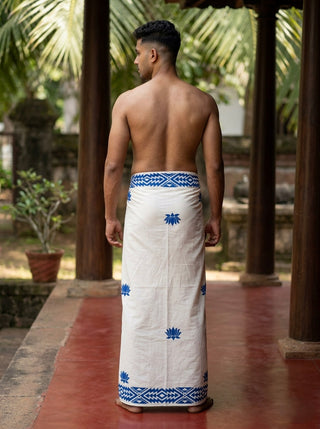 A man stands barefoot on a red floor, facing away and outdoors among trees and potted plants, wearing the Jalaj Dhoti—a traditional white cotton mundu accented with blue patterns and a handcrafted lotus motif. He is shirtless.