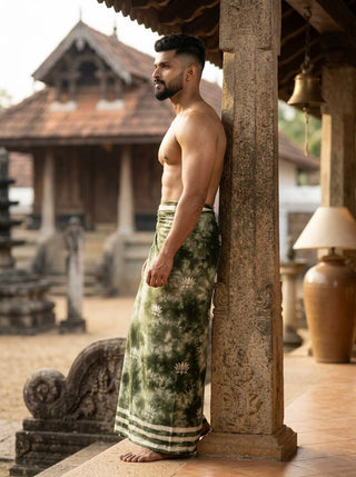 A man wearing a green Lotus Shibori Dhoti stands barefoot, leaning against a stone pillar in a courtyard with traditional architecture in the background.
