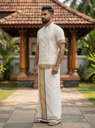 A man stands outdoors, surrounded by greenery and in front of a traditional tiled-roof structure, wearing the Bindu Heritage Shirt—a cream-colored kurta with a mandarin collar—paired with a matching mundu featuring gold detailing.