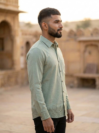 A man with a beard and short hair stands in profile outdoors against historic architecture, wearing the Madera shirt—a light green, breathable cotton shirt with subtle patterns at the bottom.