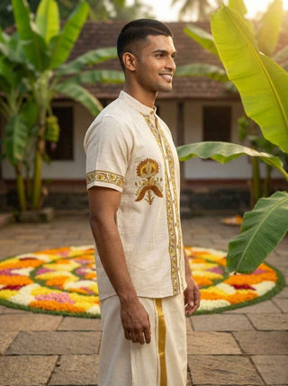 Wearing the Arosh Shirt in traditional cream and gold with ethnic styling, a man stands outdoors, smiling. Behind him is a vibrant floral rangoli on the ground and lush green banana plants.