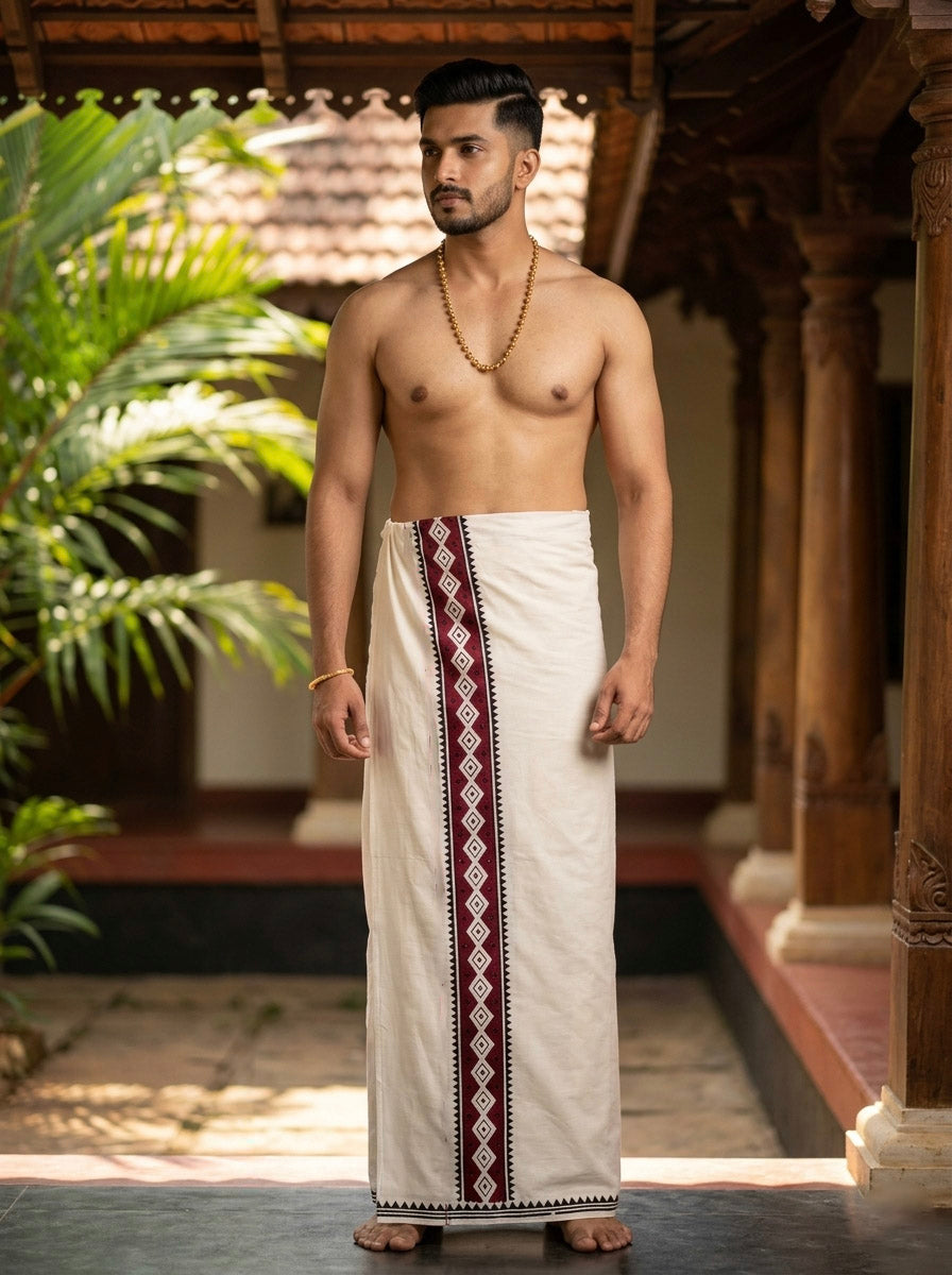A man stands barefoot in a traditional setting, wearing a white Varakha Dhoti with a maroon and black border, expertly hand block printed on premium cotton. He is shirtless, accessorized with a simple gold chain and bracelet, and looks to his right.