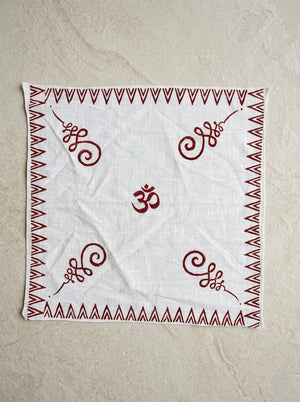 The Rudra Bandana, a white cotton accessory, showcases a bold red Om symbol at its center. Intricate Indian block print patterns adorn each corner, while a striking red zigzag border frames the design. The bandana is displayed flat against a beige textured surface.