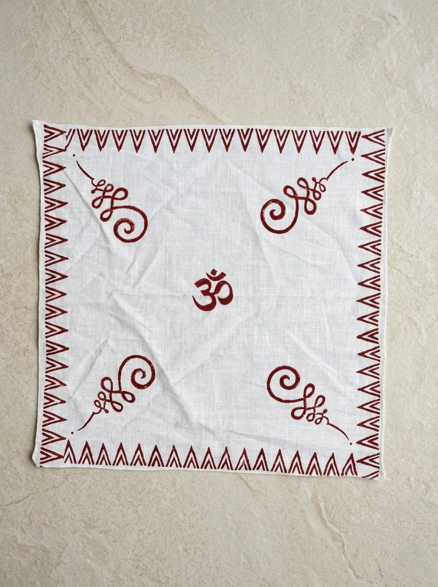 The Rudra Bandana, a white cotton accessory, showcases a bold red Om symbol at its center. Intricate Indian block print patterns adorn each corner, while a striking red zigzag border frames the design. The bandana is displayed flat against a beige textured surface.