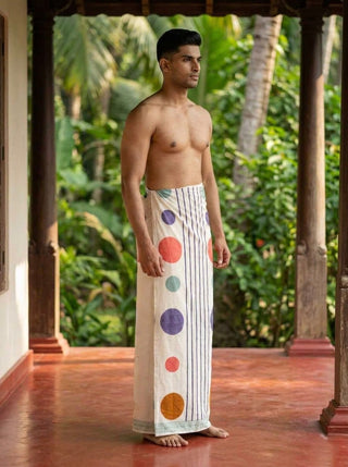 A man stands barefoot and shirtless on a veranda, surrounded by lush green plants and trees, wearing the Naaro Dhoti—a crisp white unisex cotton dhoti adorned with colorful polka dots and stripes. Reminiscent of traditional South Indian mundu styles, the Naaro Dhoti perfectly blends relaxed resort beachwear with timeless elegance.