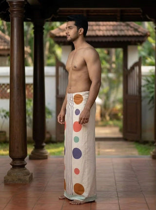 A man stands barefoot on a tiled floor, wearing the Naaro Dhoti—a traditional cream-colored South Indian garment adorned with colorful dots and stripes—with a wooden structure and greenery in the background.