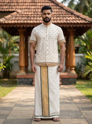 A man stands outdoors on a stone path wearing a Natural Bindu Heritage Shirt featuring patterns and a mandarin collar, paired with a traditional Indian dhoti. The background includes a tiled roof structure and lush greenery.