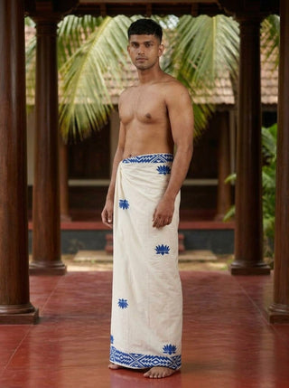 A man stands barefoot on a red floor, wearing the Jalaj Dhoti—a cream-colored traditional wrap-around mundu featuring blue patterns. He is shirtless, with wooden pillars, green palm leaves, and a tiled roof visible in the background.