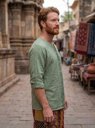 A man with red hair and a beard, wearing the Noma Shipibo Kurta made from green double gauze cotton and patterned pants, stands in profile on a stone street lined with market stalls and historic buildings.