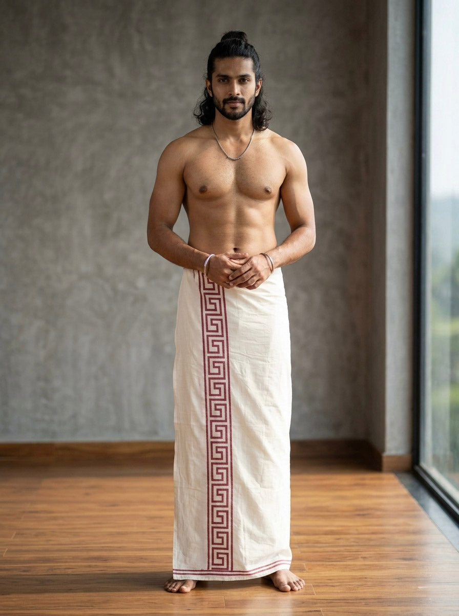 A man with long hair stands indoors, barefoot and shirtless, modeling the Rivan Dhoti—a handcrafted ethnic cotton dhoti featuring a striking red tribal border print. His hands are loosely clasped as he poses in front of a large window and gray wall.