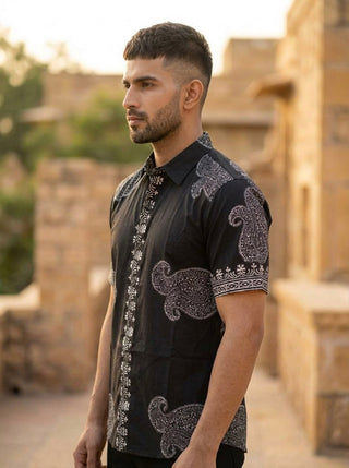 A man with short hair and a beard wears the Obsid Shirt, featuring black cotton fabric with white paisley patterns, standing outdoors near tan stone buildings and looking to the side.