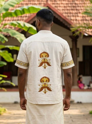 A man stands outdoors facing away, showcasing the refined ethnic styling of the Arosh Shirt—a cream-colored hand block printed shirt with floral motifs on the back—paired with a matching dhoti, set against the backdrop of a tiled-roof house and lush greenery.