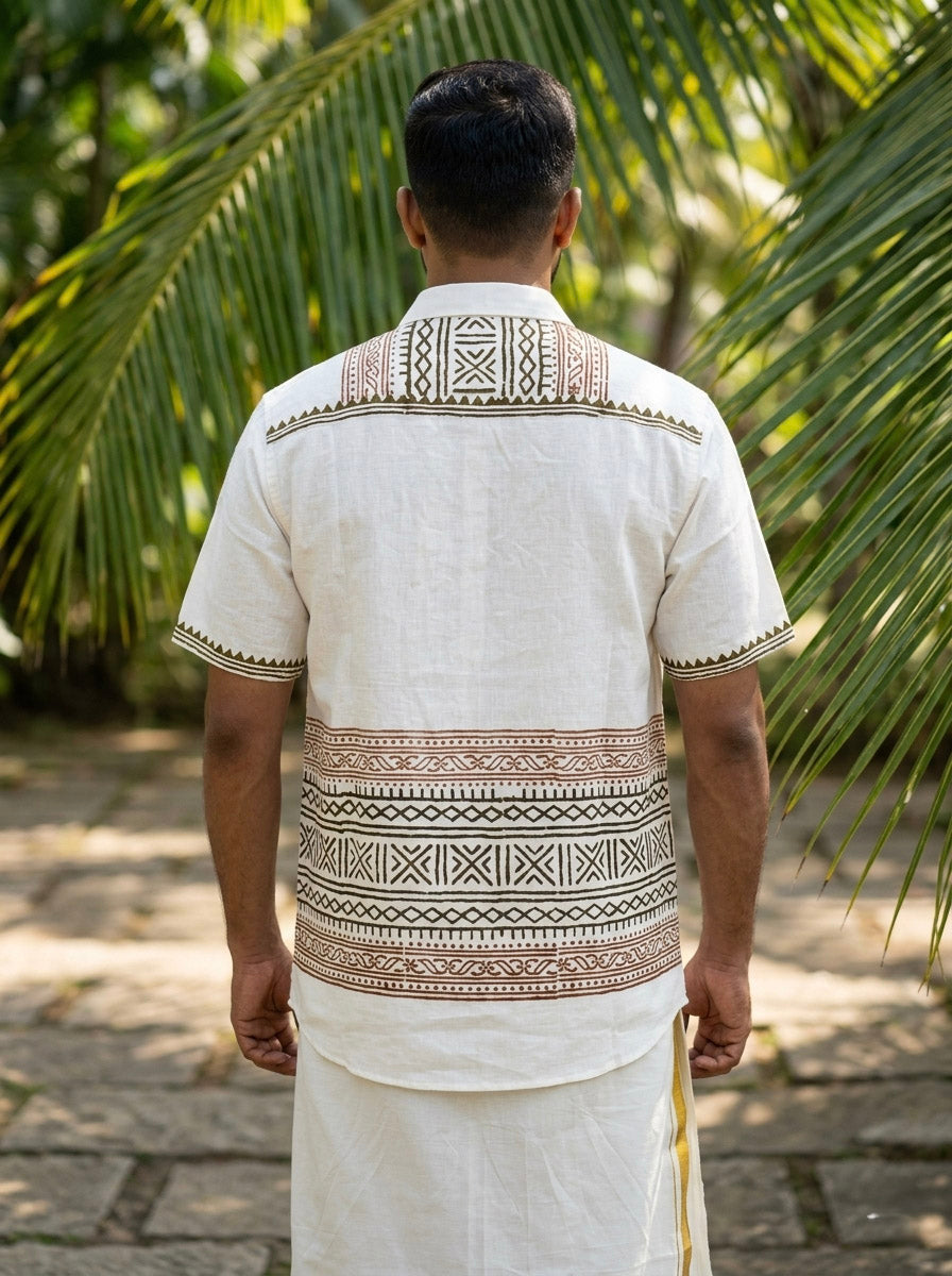A person stands outdoors with their back to the camera, wearing the white Toran Shirt and traditional attire featuring intricate brown and black patterns. Palm leaves frame the scene.