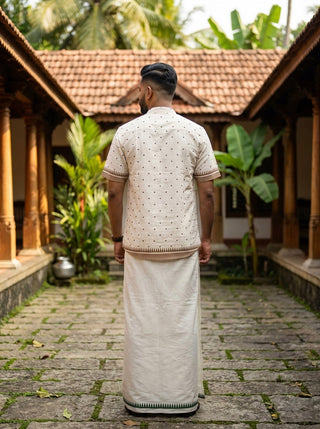 A man stands outdoors with his back to the camera, dressed in a cream-colored traditional Kerala outfit that includes the Bindu Heritage Shirt—featuring a handcrafted-inspired design and mandarin collar—paired with a mundu, surrounded by plants and tiled-roof buildings.