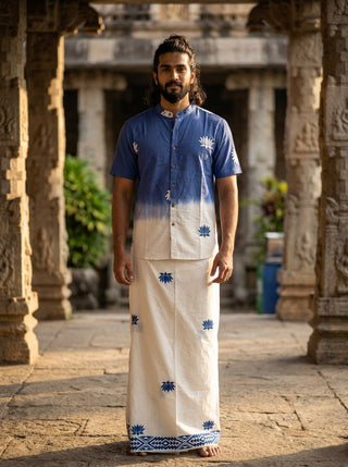 A man stands outdoors wearing the blue Sarovar Shirt, featuring intricate floral and lotus motif patterns in an ombre design, paired with a matching white traditional dhoti. Stone pillars and greenery are visible in the background.