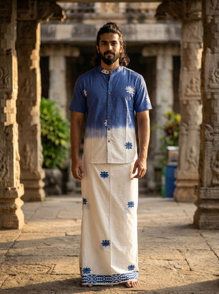 A man stands barefoot on stone pavement, wearing a blue and white short-sleeve shirt and the Jalaj Dhoti—a traditional wrap-around mundu adorned with blue patterns—in a temple-like setting with stone pillars and greenery in the background.