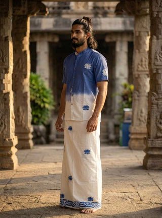 A man stands barefoot in a traditional stone courtyard, wearing the Sarovar Shirt—a blue-to-ivory ombre top with delicate lotus motifs—paired with a matching white dhoti adorned with blue lotus prints.