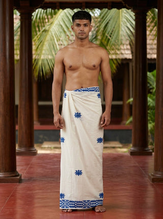 A man stands barefoot on a red floor, wearing the Jalaj Dhoti—an elegant white and blue cotton wrap adorned with handcrafted lotus motifs. He is shirtless, posing indoors amid wooden pillars and lush green palm leaves in the background.