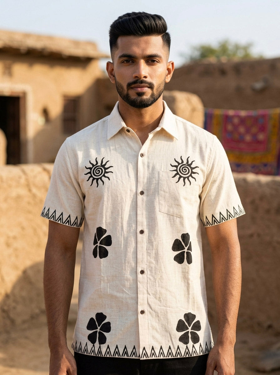 A man with short dark hair and a beard wears the Kovah Sun Shirt in Natural, featuring black geometric and floral prints, while standing outdoors in front of rustic mud buildings and colorful fabric.