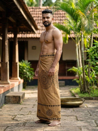 A man stands outdoors in a traditional Indian courtyard, shirtless and wearing the Kavu Dhoti featuring a heritage-inspired geometric block print. Palm trees and tiled roof buildings are in the background.