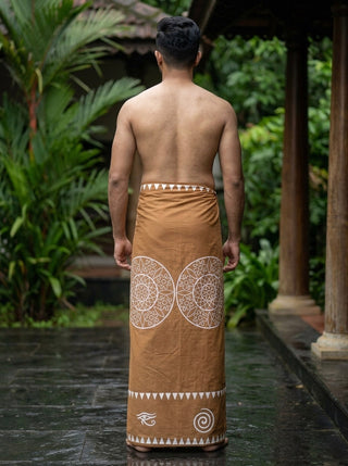 A man stands barefoot on wet stone, facing away, dressed in the Kavu Dhoti—a brown traditional garment featuring subtle mandala motifs and white patterns. Lush green plants and wooden pillars are visible in the background.