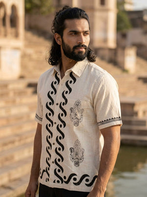 A man with long dark hair and a beard wears the cream-colored Elio Shirt featuring black artisan patterns, standing near stone steps by the water in sunlight.
