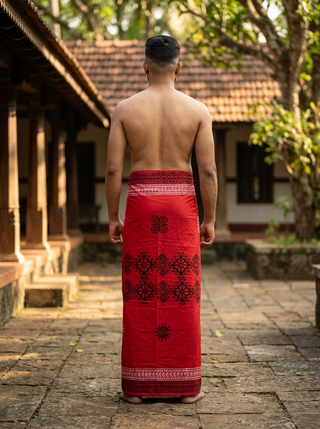 A man stands barefoot, facing away, wearing the Ramya Dhoti—crafted from breathable, lightweight fabric with a hand block print—in a traditional outdoor courtyard with trees and tiled roofs in the background. He is shirtless as sunlight filters through the scene.