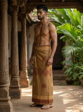 A man stands barefoot in a traditional courtyard, wearing the Ramya Dhoti—a brown hand block print garment crafted from breathable, lightweight fabric—with no shirt, surrounded by green plants and wooden pillars in the background.