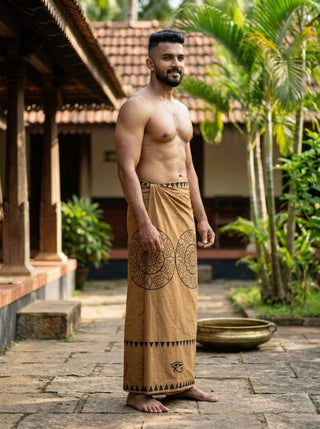 A smiling man stands barefoot on a stone path, wearing the Kavu Dhoti, which features heritage-inspired geometric block print patterns. He is shirtless, surrounded by tropical plants and old buildings.
