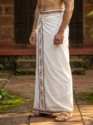 A person wearing the Mehfil Dhoti, featuring traditional white cotton fabric with brown geometric border patterns, stands outdoors on a stone path, adding an ethnic touch to the scene amidst lush greenery and a rustic building in the background.