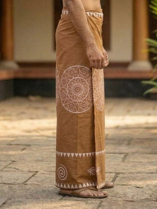 Wearing the Kavu Dhoti, a brown, ankle-length garment featuring white mandala motifs and geometric block print designs, a person stands sideways outdoors in sandals. The background reveals a paved stone area and blurred architectural elements.