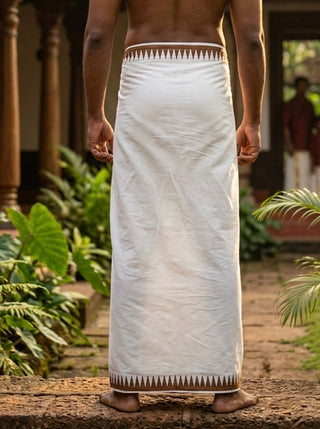 A person stands barefoot outdoors, dressed in the Mehfil Dhoti—an elegant white cotton garment accented with a patterned border—viewed from the back. Green plants and a historic building appear in the background, lending an ethnic touch to the scene.