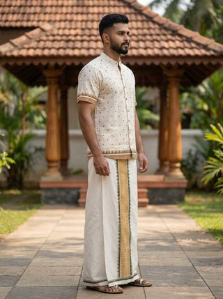 A man stands outdoors wearing the Bindu Heritage Shirt—a handcrafted-inspired cream-colored shirt with a mandarin collar—paired with a traditional mundu featuring golden borders. The tiled-roof pavilion and lush greenery in the background evoke an air of timeless elegance.