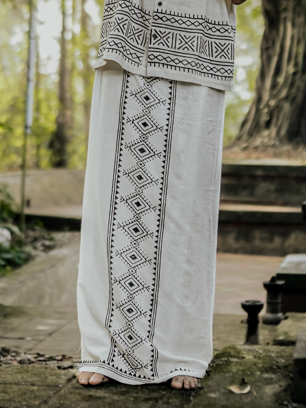 A person stands barefoot outdoors, wearing the Kangura Dhoti—a white garment featuring black geometric patterns along the front and hem. The lower half of the body is visible, showcasing the intricate details of this traditional Rajasthani handblock print.