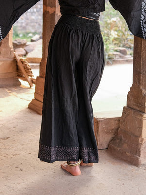 Back view of black Medusa palazzo pants between sandstone pillars, smocked waist and grey block print border at both hems