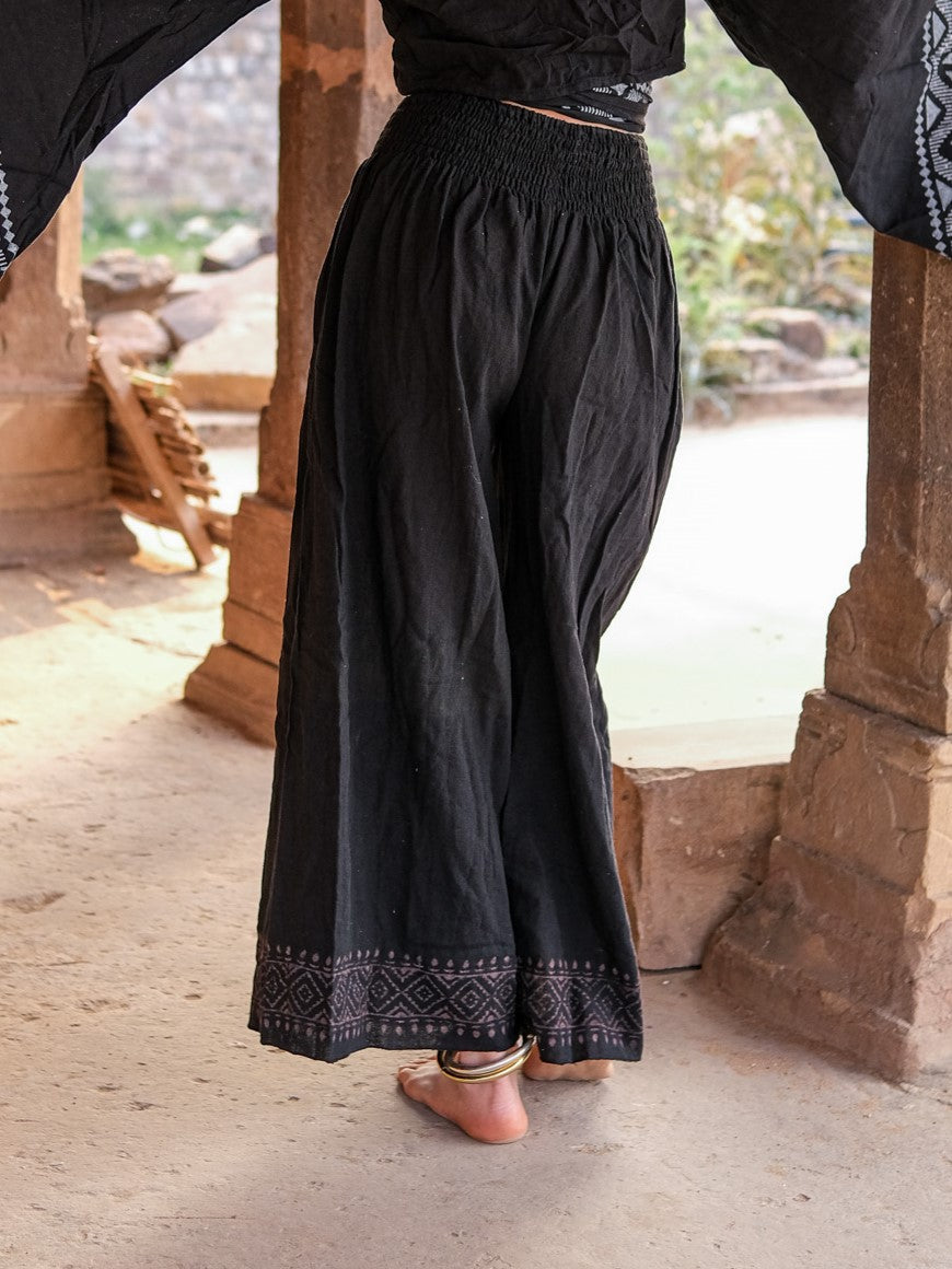 Back view of black Medusa palazzo pants between sandstone pillars, smocked waist and grey block print border at both hems