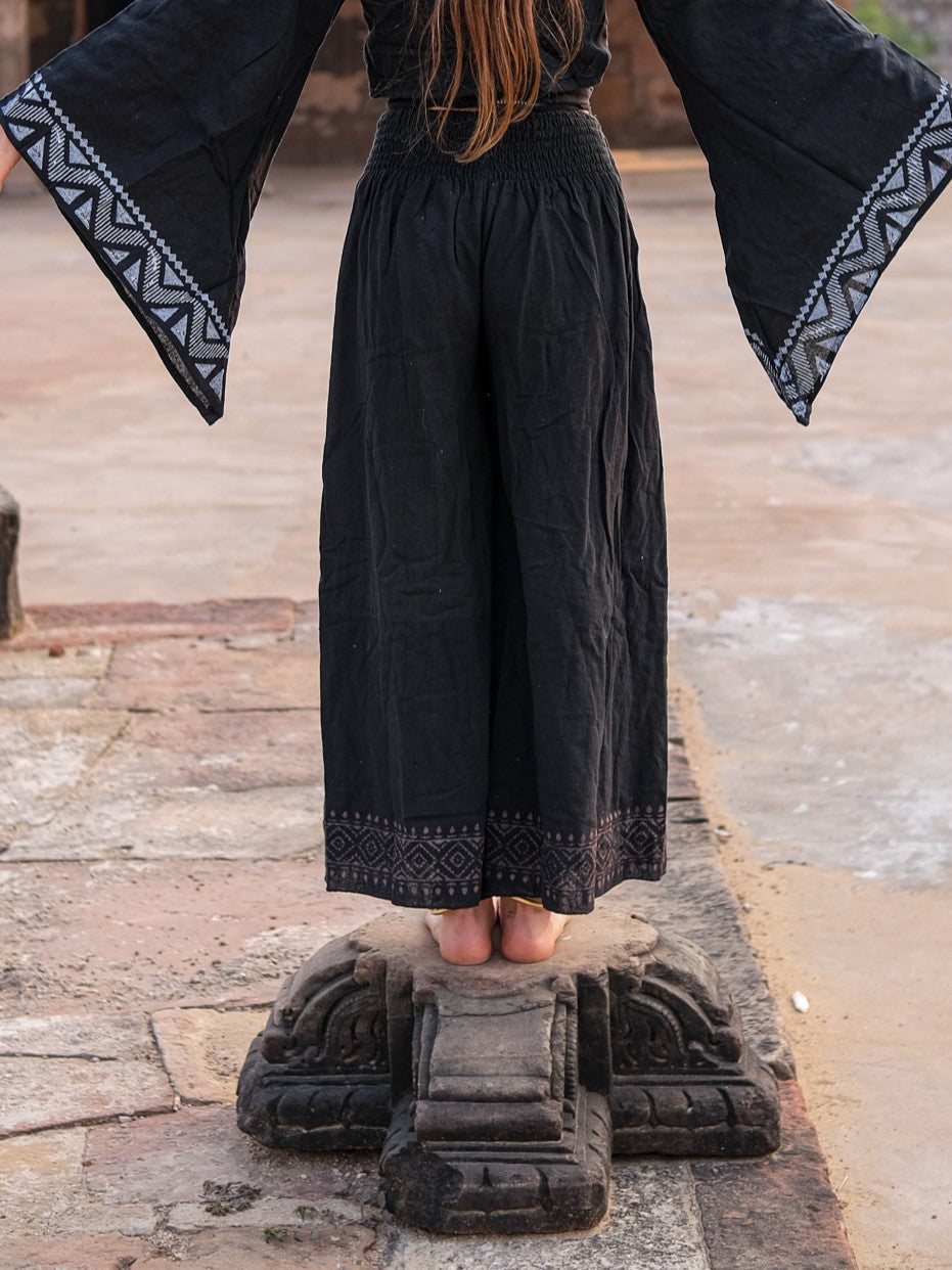 Model from behind in black Medusa palazzo pants and a black printed bell sleeve top, standing on a carved stone pedestal