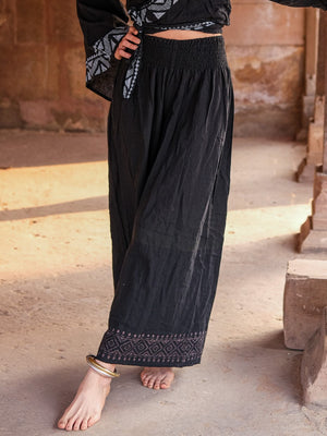 Black Medusa palazzo pants with a grey printed hem border, worn with a black patterned kimono top on a sandstone terrace