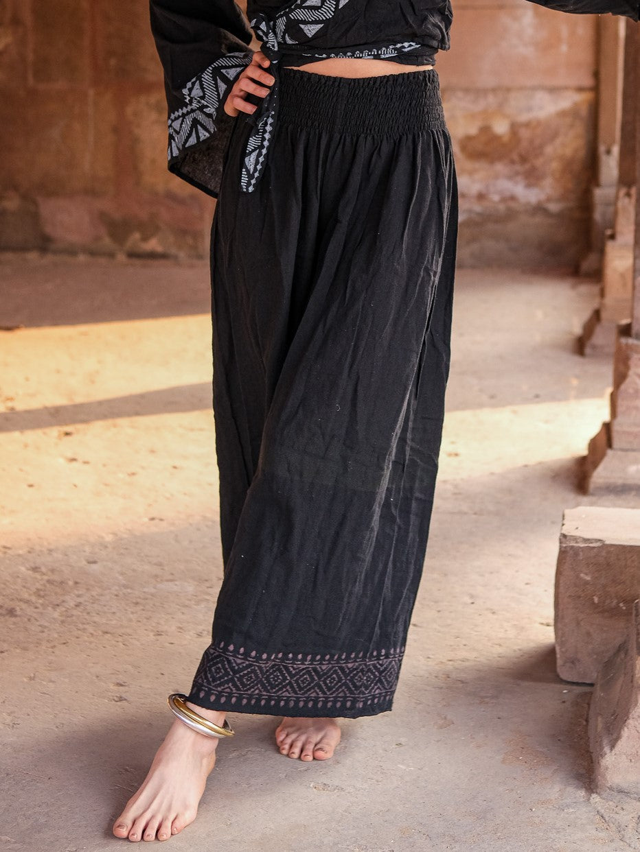 Black Medusa palazzo pants with a grey printed hem border, worn with a black patterned kimono top on a sandstone terrace