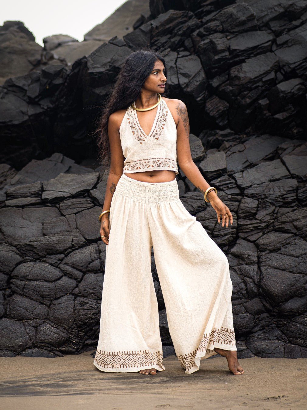 Model in natural Medusa palazzo pants and a cream embroidered halter crop top, standing before dark volcanic rocks