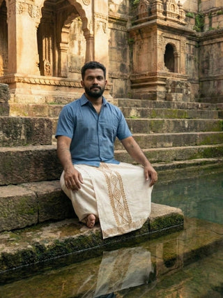 Man seated on temple steps by still water, wearing a blue shirt with the Natural Ory Dhoti in hand-printed cotton
