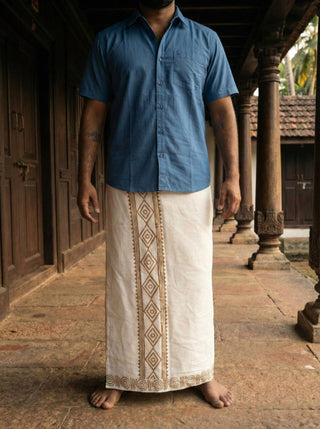 Man in a blue shirt and Natural Ory Dhoti with brown border print, standing in a carved wooden corridor